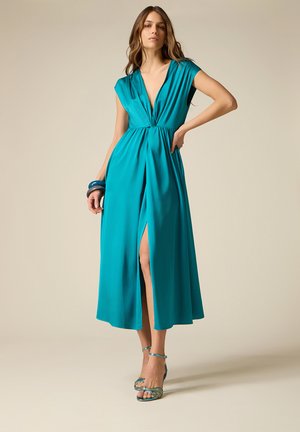 Kvinde iført en teal ærmeløs midi-kjole med dyb V-hals og frontslids, kombineret med teal stropper på hælene og blå armbånd på venstre håndled.