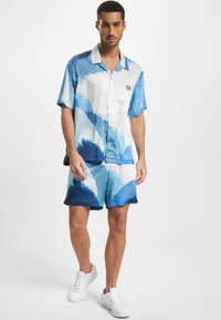 Souprava krátký rukáv, košile s límečkem a šortky v bílé a modré barvě s vlnkovými vzory. Látka vypadá lesklá a hladká. Košile je opatřena logem.
