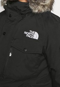Veste isolante noire avec capuche doublée de fourrure, poche à rabat, fermeture éclair et logo "The North Face" brodé en blanc sur la poitrine.