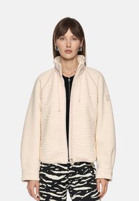 Light beige jacket with textured pattern and a high collar; features a front zipper and drawstrings. Paired with black and white patterned pants.