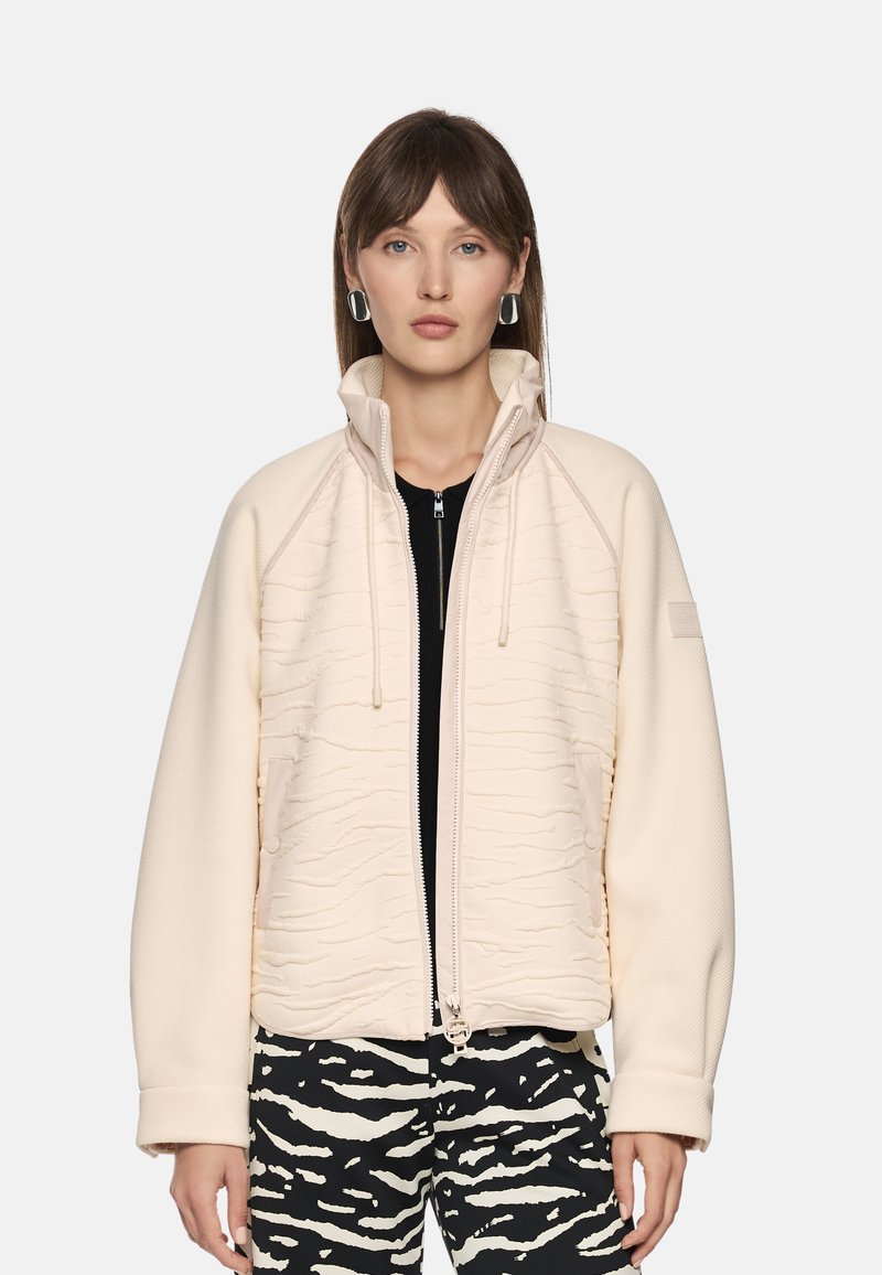 Light beige jacket with textured pattern and a high collar; features a front zipper and drawstrings. Paired with black and white patterned pants.
