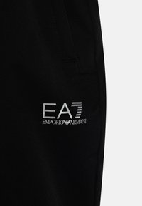 Černé sportovní kalhoty mají hladkou tkaninu s jemnou texturou. Na straně je vyšívané stříbrné logo s nápisem "EA7 EMPORIO ARMANI".