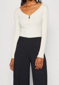 Top lungo a maniche rib a righe bianche con scollatura a V profonda e chiusura con zip frontale. Abbinato a pantaloni neri a gamba larga a vita alta.