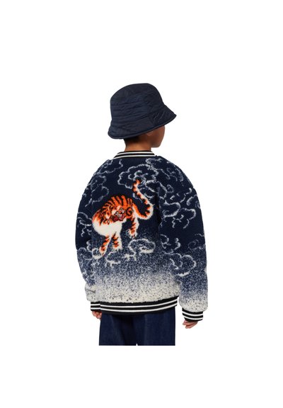 Veste bleu marine et blanche avec un design de tigre au dos, ornée de nuages et de poignets rayés. Portée avec un chapeau seau bleu marine.