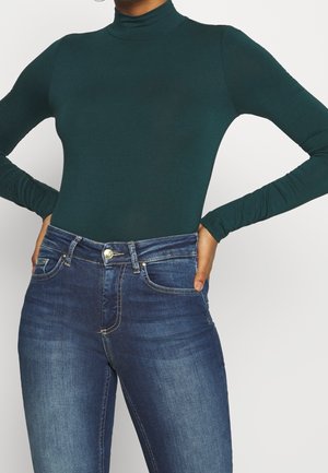 Persona con un suéter de cuello alto ajustado de color verde oscuro y jeans de mezclilla azul de cintura alta, con las manos apoyadas en las caderas.