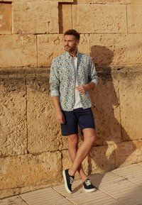 Ensemble décontracté pour homme comprenant une chemise à boutons à motif floral en blanc, bleu et jaune, associée à un short marine et des baskets noires.