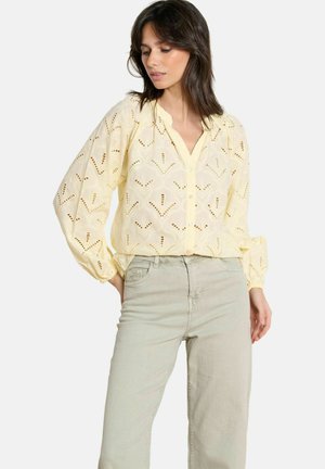 Naine, kes kannab kahvatu kollast tikanditega nööbitavat pluusi pikkade pahtelsõrmega varrukatega ja heleda halli kõrge vöökohaga pükse.