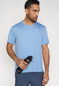 Világoskék Nike rövid ujjú póló, kerek nyakkivágással, sima textúrával, sötétkék rövidnadrággal viselve. Egy fekete vizes palackot tart, ezüst fedéllel.