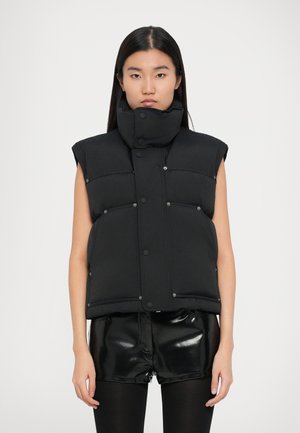 Gilet rembourré noir avec un col haut, des boutons-pression et une coupe sans manches, doté de poches avant et d'une texture lisse. Porté par-dessus un short noir brillant.