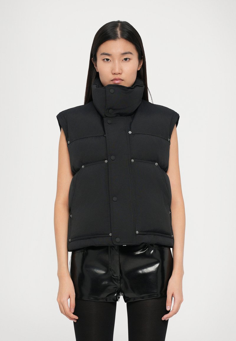 Sort polstret vest med høj krave, trykknapper og ærmeløs design, udstyret med frontlommer og en glat tekstur. Bæres over skinnende sorte shorts.