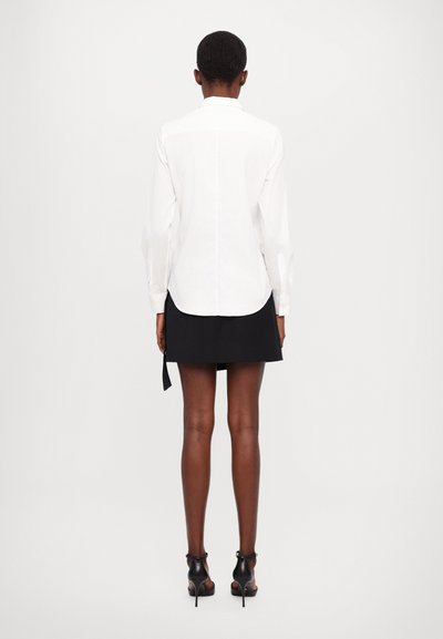 Camicia bianca a maniche lunghe con bottoni, abbinata a una gonna nera con un dettaglio di legatura laterale. Tessuto liscio, vestibilità su misura, vista da dietro.