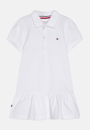 Valkoinen polo-mekko, joka on valmistettu puuvillasta. Mekossa on kauluksellinen pääntie, lyhyet hihat, nappisuljenta ja röyhelöhelma, jossa on logo-koriste.