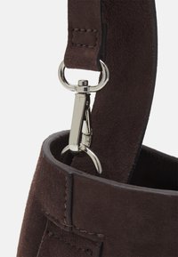 Primo piano di una tracolla in suede marrone scuro attaccata a un gancio metallico e ad un anello sul bordo di una borsa in suede abbinata.
