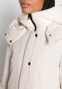 Jasno beżowa zimowa kurtka z wysokim, pikowanym kołnierzem, czarnym zamkiem błyskawicznym i zatrzaskami. Tekstura jest gładka, z lekko błyszczącym wykończeniem.