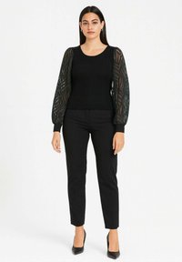 Top nero a coste con maniche nere trasparenti e decorate, abbinato a pantaloni neri aderenti e tacchi alti neri. Design semplice ed elegante.