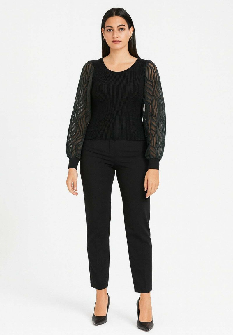 Top nero a coste con maniche nere trasparenti e decorate, abbinato a pantaloni neri aderenti e tacchi alti neri. Design semplice ed elegante.