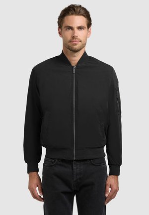 Mand iført en sort bomberjakke med lynlås, ribstrikkede manchetter og krave, stående foran en ensartet grå baggrund.