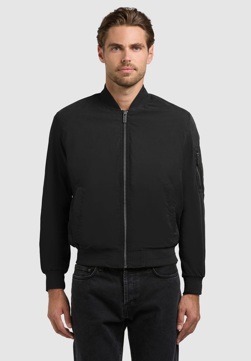 Homme portant une veste bomber noire zippée avec des poignets et un col côtelés, debout devant un fond gris uni.