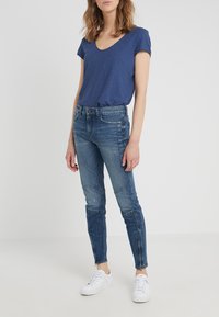 Modrá trikó s výstřihem do V spárované s potrhanými modrými skinny džíny s detaily kapes a vybledlým vzhledem, doplněné bílými teniskami.