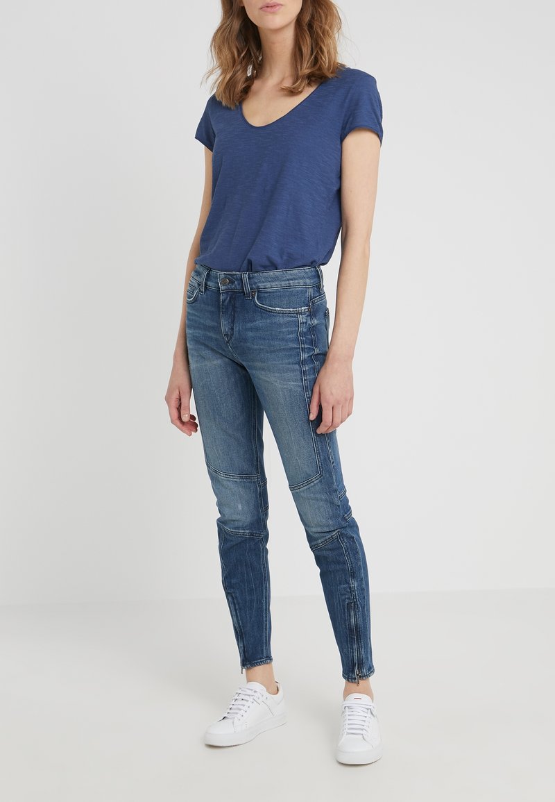 Modrá trikó s výstřihem do V spárované s potrhanými modrými skinny džíny s detaily kapes a vybledlým vzhledem, doplněné bílými teniskami.