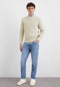 Cremefarbener, gerippter Strickpullover, hellblauses Hemd darunter, blaue Jeans und beige Sneakers mit weißen Akzenten. Einfaches, lässiges Outfit-Design.