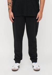 Zwarte trainingsbroek van zacht materiaal, met een ontspannen pasvorm, elastische tailleband en boorden. Gepaard met witte sneakers met zwarte strepen.