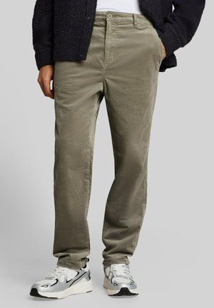 Mann trägt hellbraune Cordhose, weiße und graue Turnschuhe und eine schwarze Jacke mit einer Hand in der Tasche.