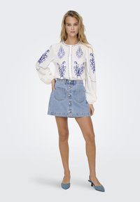 White blouse with blue embroidered patterns, paired with a light denim mini skirt featuring front buttons and two back pockets. Blue heels.