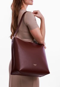 Brown leather tote bag with a smooth texture, featuring a gold logo and a wide shoulder strap. The bag has a minimalist design.