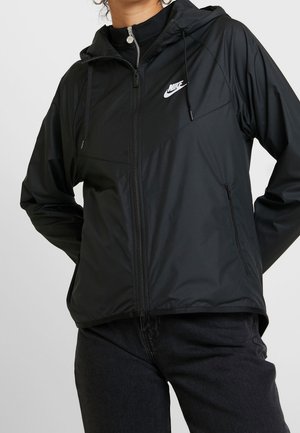 Mujer con chaqueta negra con capucha y cremallera de Nike y pantalones negros, de pie con las manos detrás de la espalda sobre fondo liso.