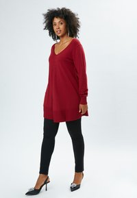 Rote Strick-Tunika mit V-Ausschnitt und dezentem Plissee-Detail; kombiniert mit schwarzen Skinny-Jeans und schwarzen Slingback-Absatzschuhen mit dekorativen Akzenten.