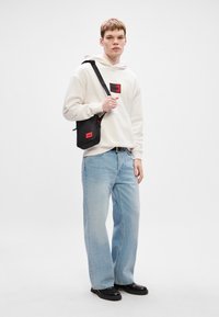 Μαύρη τσάντα χιαστί με κόκκινες λεπτομέρειες, φορεμένη από ένα άτομο με μπεζ φούτερ και ανοιχτόχρωμα τζιν. Απλό σχέδιο με ρυθμιζόμενο λουρί.