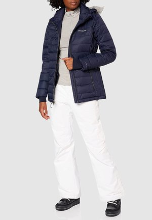 Marineblaues Puffer-Jackett mit durchgehendem Reißverschluss, mit Fell gefütterter Kapuze und gestepptem Design. Getragen mit einem hellgrauen Oberteil und weißen Schneehosen. Schwarze Schuhe.
