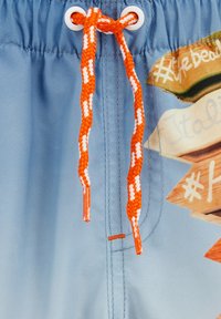 Modré plavky s pletenou oranžovou a bielou šnúrkou, so kovovými očkami. V pozadí sú tlačené smerové značky.