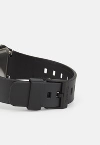 Montre numérique noire avec un bracelet en caoutchouc, boîtier rectangulaire, bande texturée et une boucle. Présente un marquage sur le bracelet et des trous ajustables.