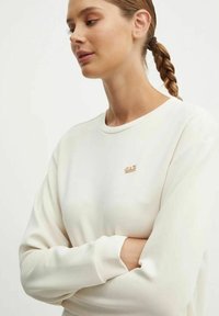 Maglione chiaro in crema con maniche lunghe e scollatura rotonda. Presenta un piccolo logo metallico sul petto. Tessuto dalla consistenza liscia.