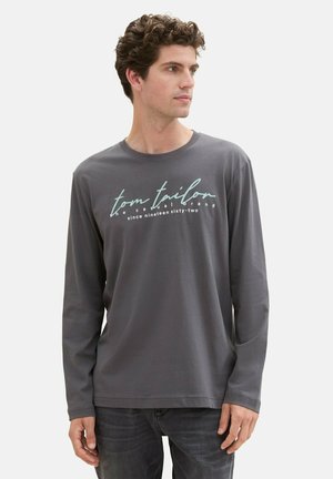 Camiseta de manga larga gris, hecha de una tela suave, con texto impreso en blanco y aguamarina: "tom tailford" y "desde mil novecientos sesenta y dos."