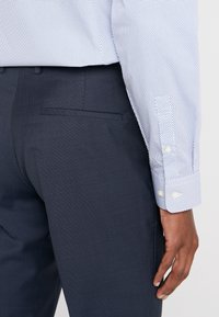 Weißes Hemd mit blauen Poi-Punkten, mit geknöpften Manschetten; kombiniert mit dunklen, strukturierten Hosen. Nahaufnahme, die sich auf die Details der Manschette und Tasche konzentriert.