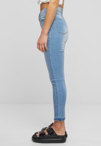 Világoskék, magas derekú, skinny farmernadrág sima denim textúrával és rojtos szegéllyel, fekete szandálokkal párosítva, széles pántos dizájnnal.