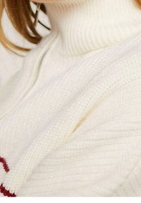 Donna che indossa un pullover a collo alto a coste di colore crema, con un dettaglio rosso parziale visibile vicino al bordo, capelli che poggiano sulla spalla.