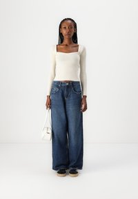 Haut à manches longues blanc côtelé associé à un jean en denim bleu foncé, ample. Le mannequin tient un sac à main blanc ; chaussures noires avec semelles beige.