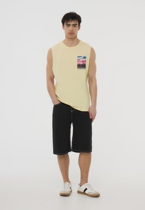 Maglietta gialla senza maniche unisex con una tasca grafica, abbinata a pantaloni corti neri fino al ginocchio con cuciture bianche e scarpe da ginnastica bianche.