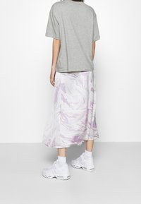 Osoba ubrana w szary T-shirt z krótkim rękawem, midi spódnicę w białe i lawendowe marmurkowe wzory, białe skarpetki i białe trampki, stojąca na tle jednolitego tła.