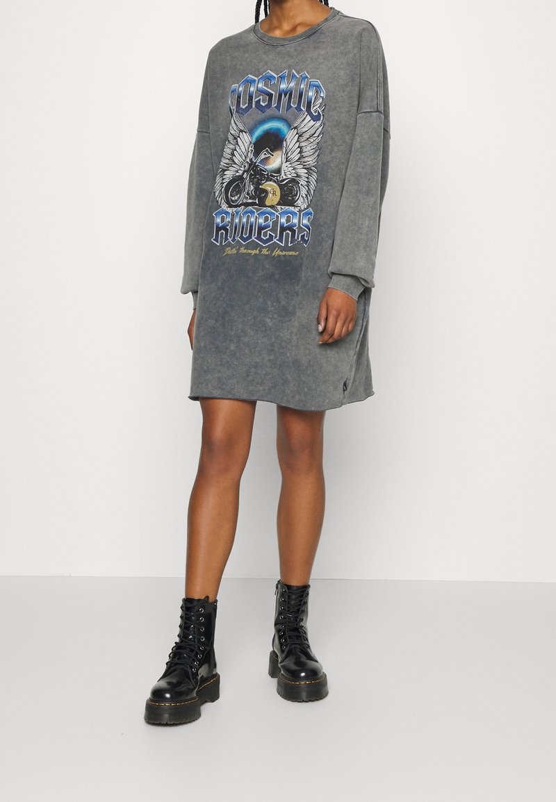 Femme portant une robe longue ample grise à manches longues avec l'inscription « Cosmic Riders » et un graphisme de moto ailée, associée à des bottines noires à lacets.