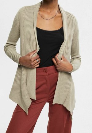 Frau trägt einen beige gerippten cardigan mit offenem Vorderteil über einem schwarzen Oberteil und rostfarbenen, hoch taillierten Hosen, hält die Kanten des Cardigans fest.