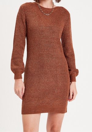 Femme portant une robe pull en maille de couleur rouille avec des manches longues et un collier chaîne argenté, posant devant un fond blanc uni.