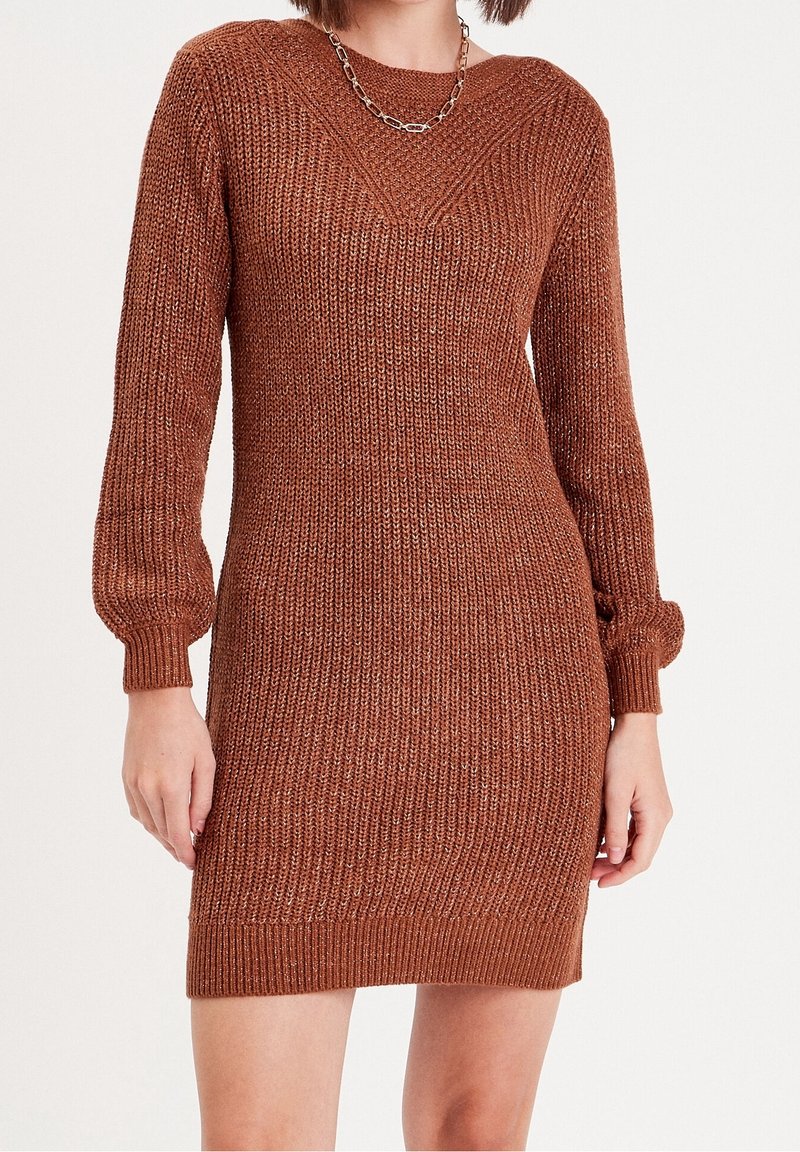 Femme portant une robe pull en maille de couleur rouille avec des manches longues et un collier chaîne argenté, posant devant un fond blanc uni.