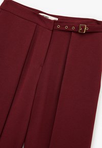 Burgunderfarbene Hose aus glattem Stoff, mit einem gefältelten Vorderteil, gestepptem Bund und einem goldfarbenen Schnallen-Detail am Gürtel.
