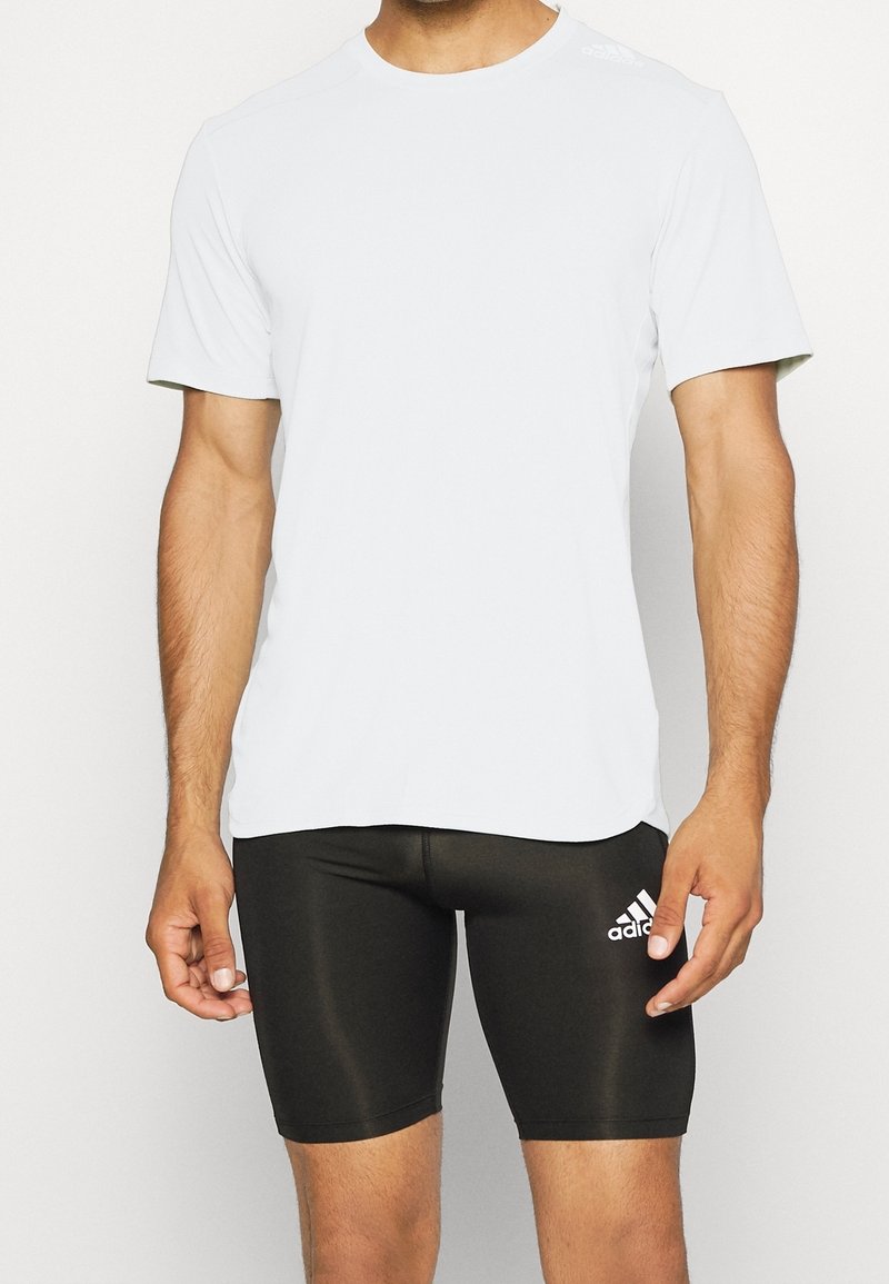 Camisa atlética blanca de manga corta con cuello redondo, combinada con pantalones cortos ceñidos negros que tienen un logo blanco en el muslo izquierdo.