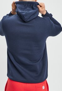 Felpa blu navy con finitura testurizzata, dotata di cappuccio con cordini e polsini a costine, abbinata a pantaloni da tuta rossi.